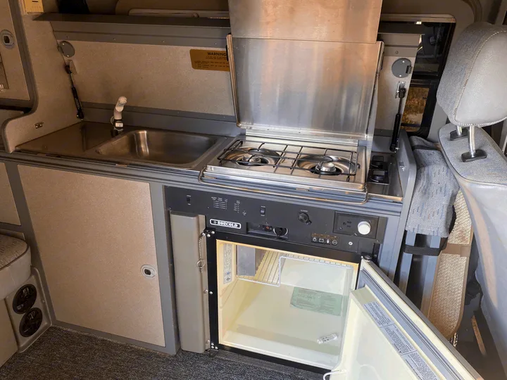 Compact RV rental kitchen featuring a stainless steel sink, two-burner stove, and open Norcold refrigerator, perfect for travel.. Volkswagen T4 Multivan Westfalia 1995