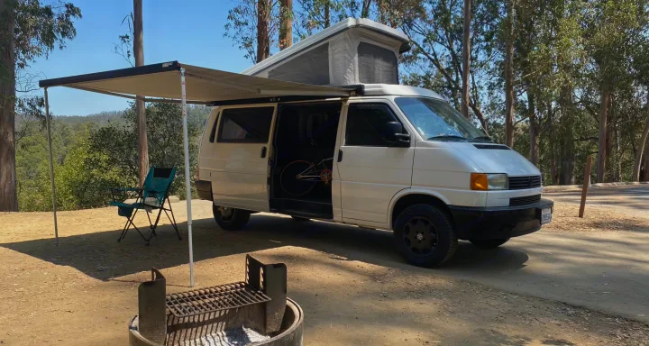 Sunny campsite: white pop-top camper van with extended awning, chair, fire pit, and bike inside. Your rv rental escape.. Volkswagen T4 Multivan Westfalia 1995