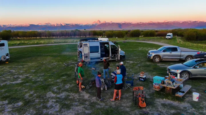 Friends by campfire at scenic RV rental campsite, with camper van, vehicles, and snow-capped mountains at sunset.. Mercedes-Benz Sprinter 2016