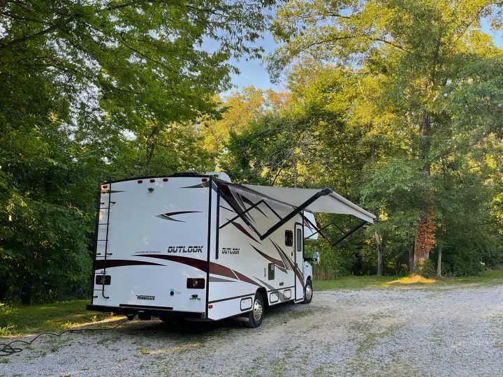 White Winnebago Outlook RV rental parked on gravel with awning extended, surrounded by green trees. Perfect for camping.. Winnebago Outlook 2020