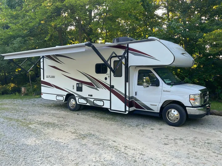This white RV rental, an Outlook model motorhome, is parked on a gravel lot with its awning extended amidst green trees, ready for camping.. Winnebago Outlook 2020