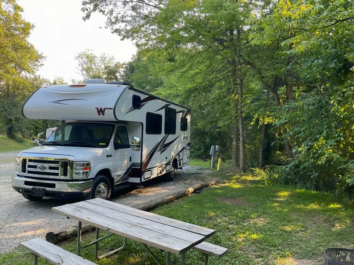 White Class C rv rental parked on gravel at a wooded campsite with a wooden picnic table, ready for outdoor adventure.. Winnebago Outlook 2020