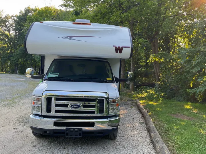 A white Ford Class C RV rental is parked on a gravel site, surrounded by green trees at a campground.. Winnebago Outlook 2020