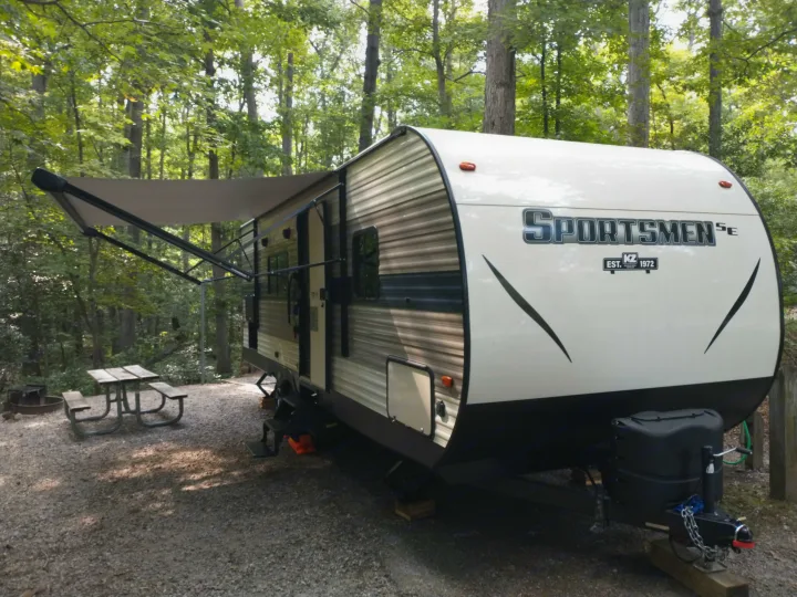 Enjoy nature with this RV rental! A travel trailer with an extended awning, picnic table, and fire pit is parked at a wooded campsite.. KZ Sportsmen 2021