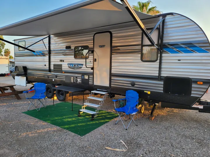 A comfortable rv rental travel trailer parked with its awning out, blue chairs, and picnic table at a sunny gravel campsite.. Forest River Salem 2021