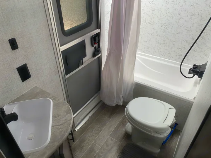 Compact RV rental bathroom: corner sink, interior door, toilet, and bathtub with curtain. Light walls, wood-look floor.. Gulf Stream Kingsport 2021
