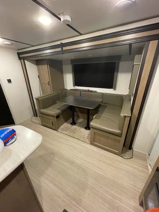 Bright RV rental interior showing a U-shaped dinette with grey cushions, a black table, light wood floor, and window.. Keystone Hideout 2021