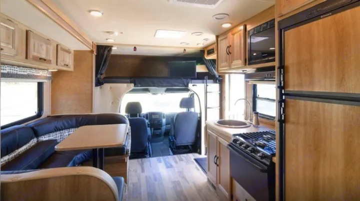 Inside a light-wood RV rental, showing the living/dining area with dark seating, a kitchen with appliances, and the driver's cab.. Coachmen Prism 2016