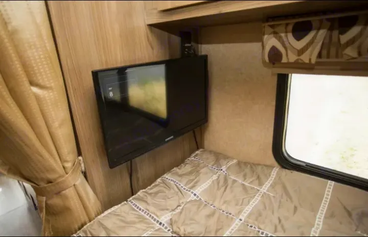 RV rental interior: a compact bedroom features a wall-mounted TV, cozy bed, window, and curtain.. Coachmen Prism 2016