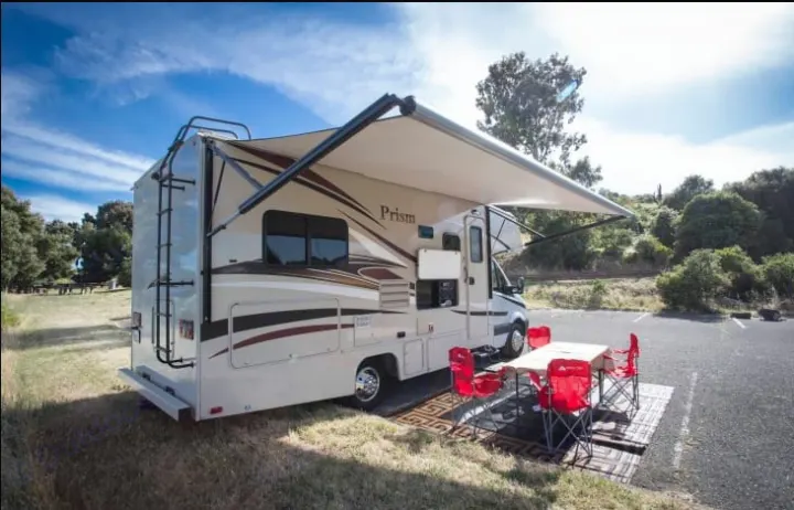 A light-colored rv rental with an extended awning, outdoor table, and red chairs is set up at a scenic campsite.. Coachmen Prism 2016
