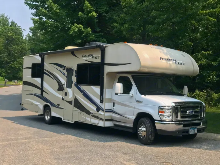 This beige & white Class C motorhome, a Freedom Elite rv rental, is parked on asphalt, surrounded by lush green trees.. Thor Motor Coach Freedom Elite 2015