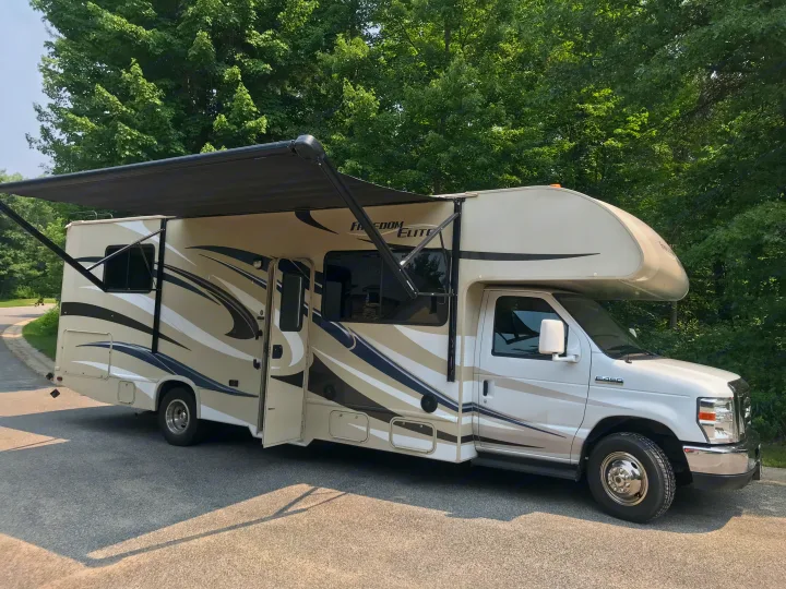 This beige Freedom Elite Class C **rv rental**, with an extended awning, is parked on an asphalt road near trees.. Thor Motor Coach Freedom Elite 2015