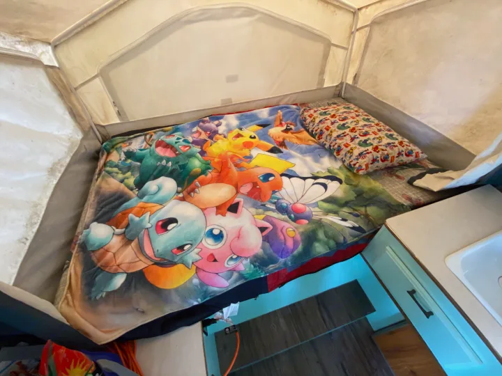 Cozy RV rental interior featuring a bed with a vibrant Pokémon blanket, Marvel pillow, and a compact kitchen area with a sink.. Forest River Rockwood Freedom 2001