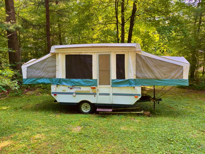 This white and green Rockwood pop-up camper RV rental is fully set up in a lush forest clearing, perfect for camping.. Forest River Rockwood Freedom 2001