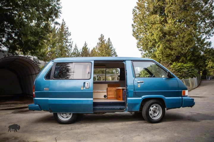 Blue camper van, resembling a Toyota, with its side door open revealing a cozy wood-finished interior. Ideal for an RV rental.. Toyota Hiace 1985
