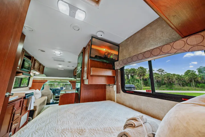 Comfortable rv rental interior with a cozy bed, mounted TV, kitchen, and scenic window view, ideal for travel.. Winnebago Spirit 2015