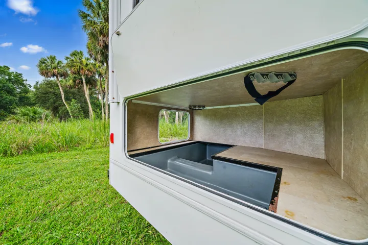 RV rental: Open cargo bay reveals gray storage bin, raised floor, ceiling hooks, and an outdoor view of lush green scenery.. Winnebago Spirit 2015