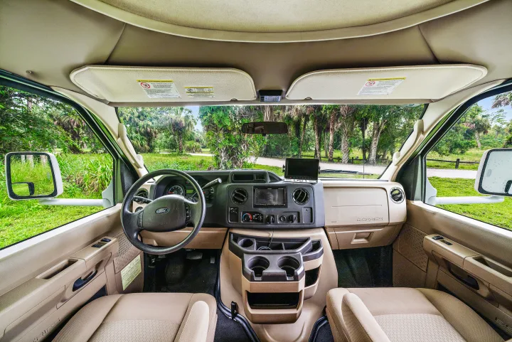 RV rental interior view: dashboard, steering wheel, and front seats, with lush forest visible through the windshield.. Winnebago Spirit 2015