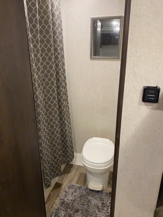 RV rental bathroom: compact toilet, gray patterned shower curtain, small mirror, shaggy gray rug, & thermostat.. Starcraft Autumn Ridge 2018