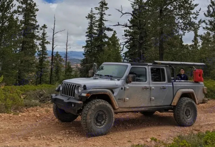 A rugged gray Jeep Gladiator pickup truck on a dirt trail in a forest, with a person in the back. Like an rv rental, perfect for adventure!. Jeep Gladiator 2020