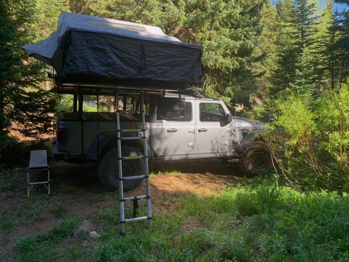 Grey Jeep Gladiator with roof-top tent set up for camping in a lush forest campsite. A modern rv rental adventure option.. Jeep Gladiator 2020