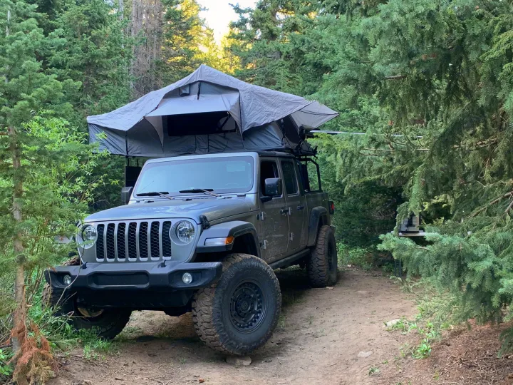 An overlanding Jeep Gladiator with a deployed rooftop tent, an alternative RV rental, parked on a dirt trail amidst a dense forest.. Jeep Gladiator 2020