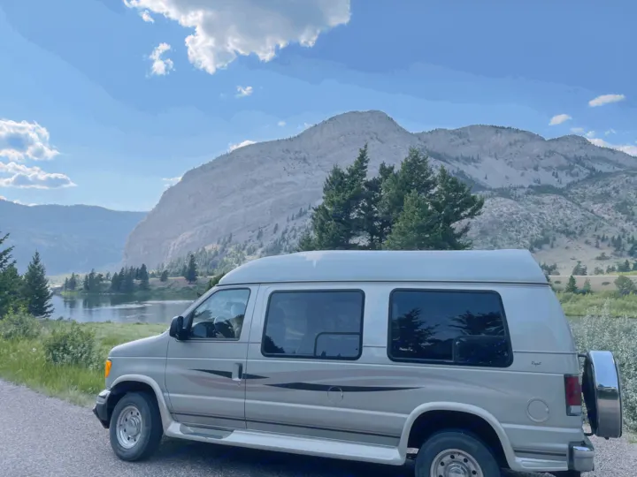 An RV rental van by a serene river, showcasing grand mountains under a blue sky. Perfect for outdoor adventures.. Ford Econoline 2006
