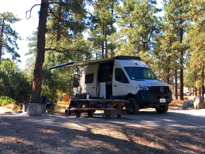 A white Mercedes-Benz Sprinter RV rental parked at a serene campsite with an extended awning among tall pines.. Winnebago Revel 2021
