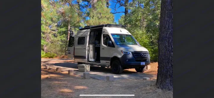 Experience the outdoors in this beige Mercedes Sprinter **rv rental** parked at a wooded campsite with its doors open.. Winnebago Revel 2021