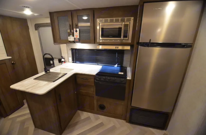 Inside an RV rental, a well-equipped kitchen with a large stainless steel refrigerator, microwave, stove, sink, and wood cabinetry.. Forest River Wildwood X-Lite 2022