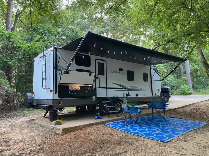 An RV rental is set up at a serene campsite with an extended awning, string lights, camping chairs, and a blue mat.. Palomino Solaire Ultra Lite 2021