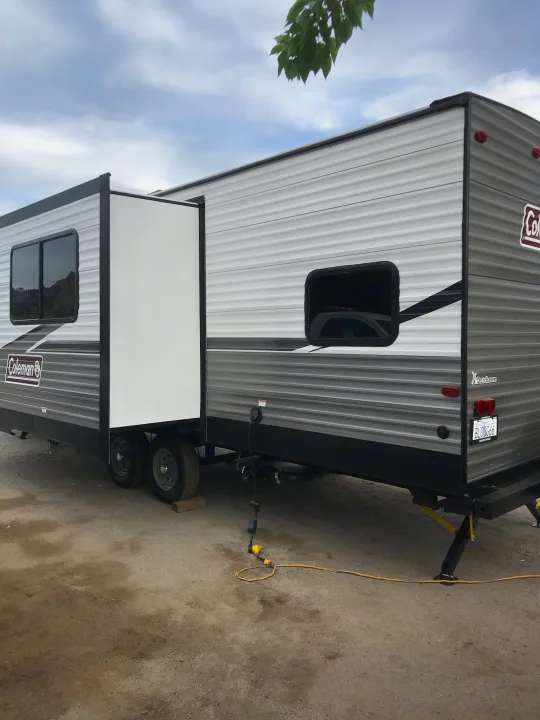 A large grey and white Coleman travel trailer with its slide-out extended, set up outdoors for an RV rental stay. Ready for camping!. Coleman 295QB 2021