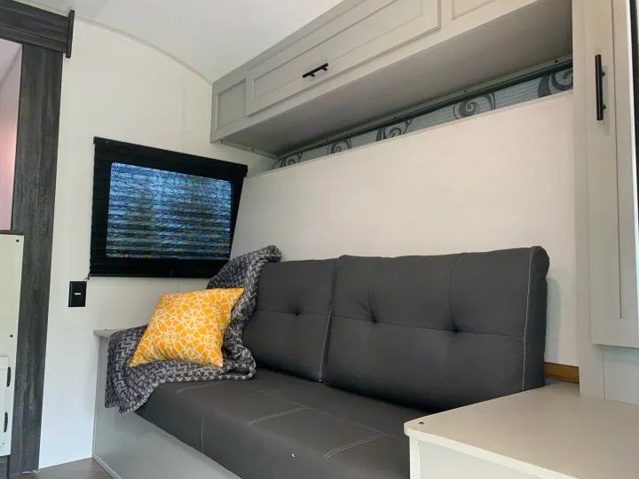 RV rental living space with dark grey futon, yellow pillow, window, and light grey overhead cabinets.. Forest River Salem 2021