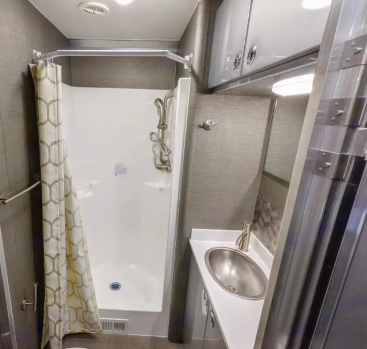 This RV rental bathroom features a compact shower with a patterned curtain, an oval sink vanity, and overhead storage.. Aluminum Trailer Company 2816 2021