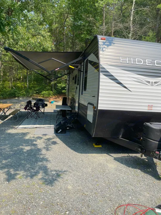 RV rental parked at a shaded campsite with an extended awning, outdoor furniture, and picnic table, ready for a cozy outdoor experience.. Keystone Hideout 2021