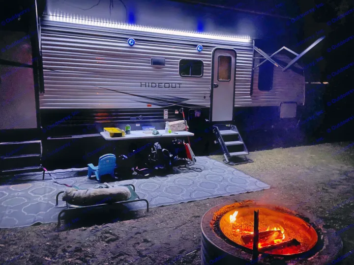 Cozy RV rental setup at night: bright trailer lights, a glowing campfire, and a dog on its bed.. Keystone Hideout 2021
