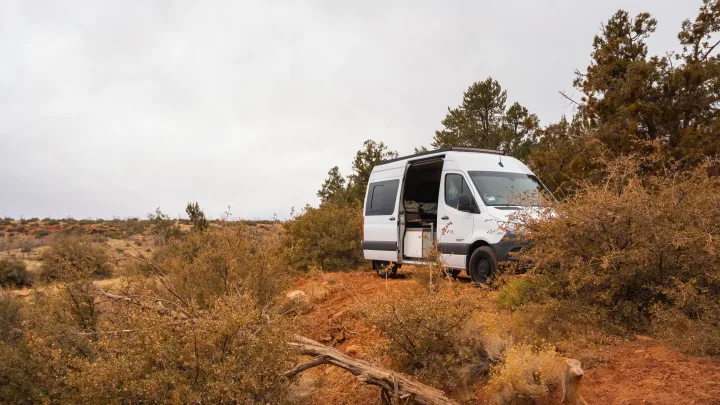 Explore nature with an rv rental. This white camper van is parked on red dirt, side door open, amidst arid bushes under a grey sky.. Mercedes-Benz Sprinter 2020
