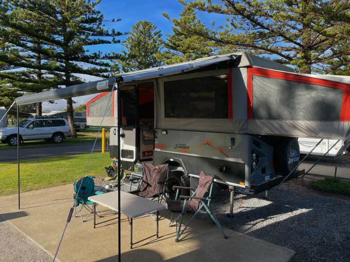 A grey & orange pop-up RV rental with extended awning, outdoor table, & chairs on a concrete slab, set amidst pine trees & green grass.. Jayco Swan Outback 2021