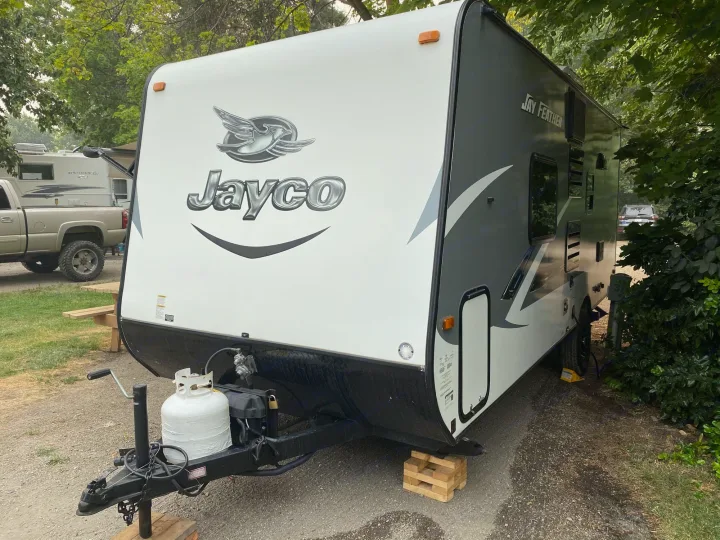 Front view of a white and gray Jayco travel trailer at a campground, ready for an RV rental adventure with its propane tank visible.. Jayco Jay Feather 2017