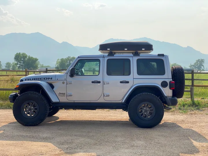 Silver Jeep Wrangler Rubicon with roof box on gravel, mountains in background. Ideal adventure vehicle, great RV rental choice.. Jeep Wrangler Unlimited Rubicon 392 2021
