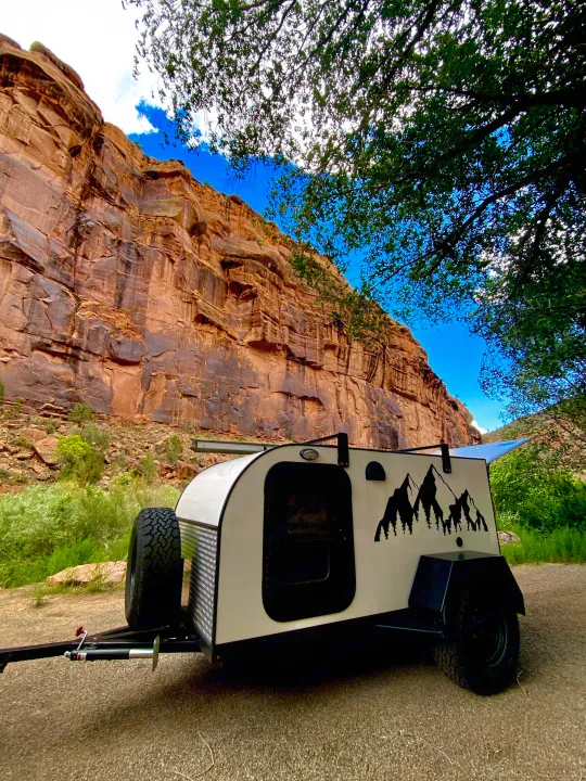 A unique teardrop camper, ideal as an RV rental, parked by a towering red rock canyon wall under a blue sky, surrounded by green nature.. Other Other 2021