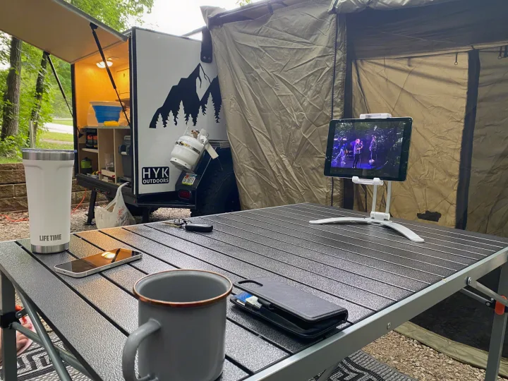 A modern RV rental campsite scene features a cozy outdoor kitchen, a durable folding table with a tablet displaying entertainment, and essentials.. Other Other 2021