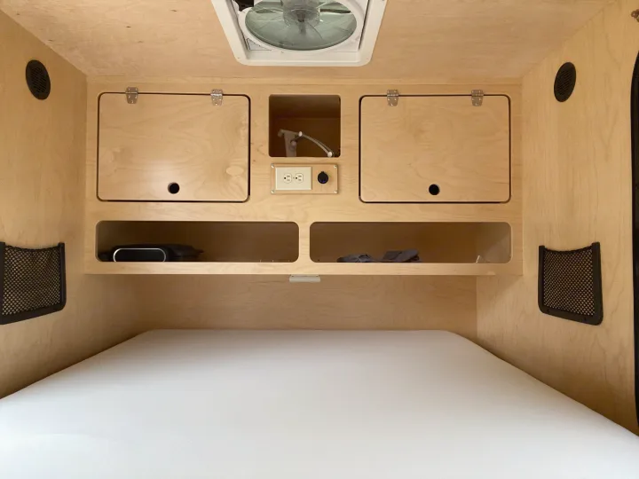 View of an rv rental's cozy interior: white bed, wooden storage, power outlets, ceiling fan, and mesh pockets.. Other Other 2021