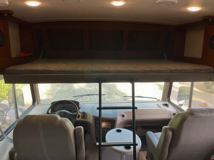 Inside an RV rental: view of the front cockpit with steering wheel and seats, plus an overhead bunk bed and ladder.. Forest River Forester 2018