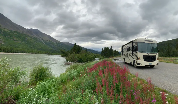 Scenic RV rental journey: a motorhome on a road beside a river with mountains, green forests, and colorful flowers under a cloudy sky.. Forest River Forester 2018
