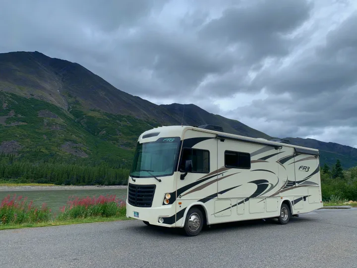 An RV rental motorhome, white with graphics, is parked by a river with green mountains and cloudy skies. Ready for adventure.. Forest River Forester 2018