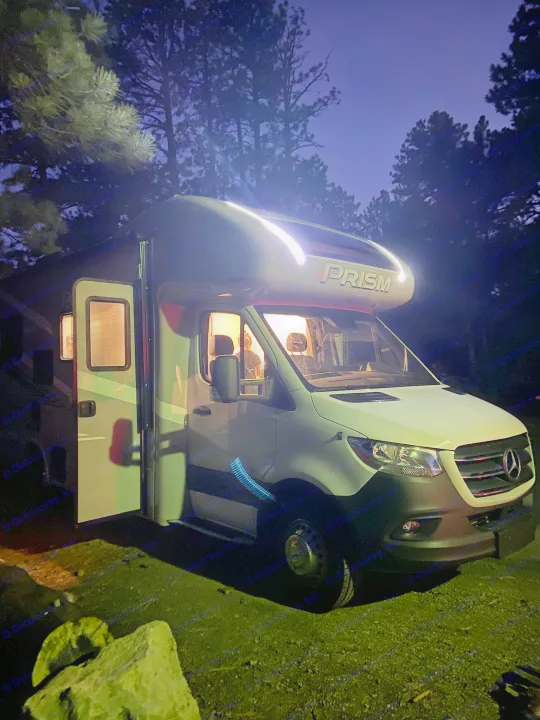 An illuminated Mercedes-Benz RV rental glows warmly at dusk, parked amidst trees with its door open for a cozy night.. Mercedes-Benz Sprinter Coachmen Prism 2021