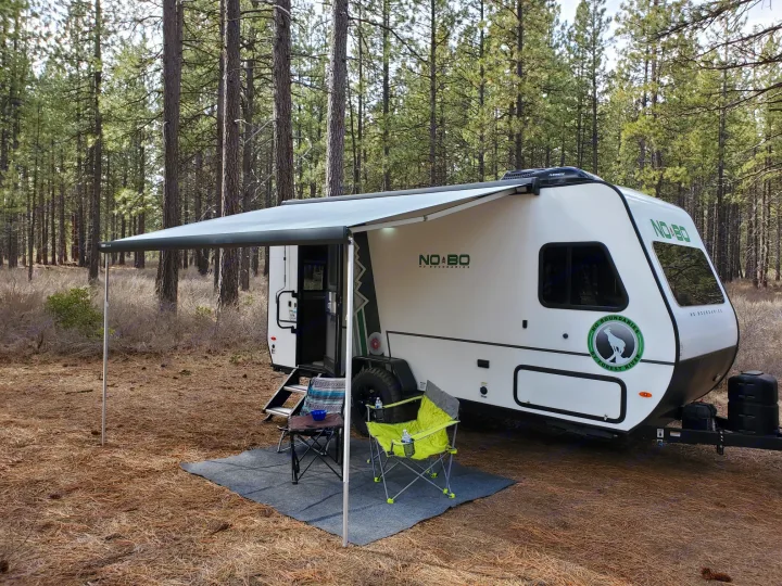 RV rental camping in a serene pine forest. A white travel trailer with awning, chairs, and table on a mat.. Forest River Other 2019