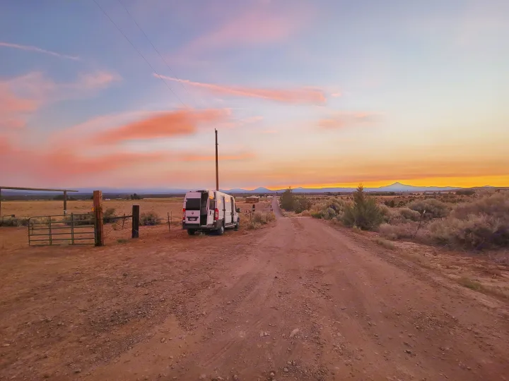 Stunning sunset paints the sky over an RV rental van on a dusty desert road, framed by distant mountains.. Dodge Promaster 2500 High Roof 2020