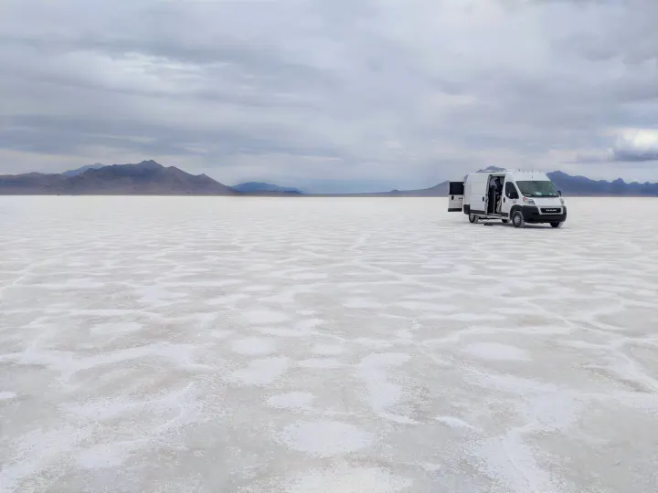 White RV rental van on vast Bonneville Salt Flats with open doors, mountains under cloudy sky.. Dodge Promaster 2500 High Roof 2020